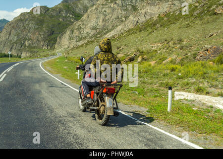 Due ragazzi senza casco ride su un vecchio rosso sovietica motocicletta sulla strada nelle montagne di Altai da un villaggio all altro, uno è vestito in Foto Stock