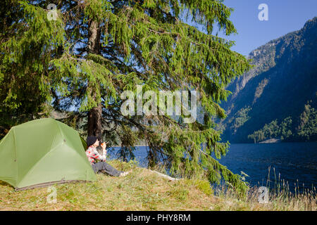 Donna traveler godendo il cappuccio del caffè o del tè seduto in tenda da campeggio su un lago sotto abete presso Scenic campeggio selvaggio in Norvegia Foto Stock