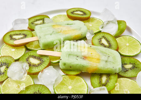 Close-up di calce viva in casa kiwi icy ghiaccioli con un pezzo di pesca in una piastra con cubetti di ghiaccio e pezzi di frutta Foto Stock