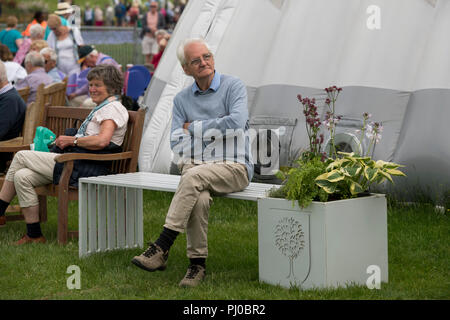 Senior uomo seduto da solo su bianco sedile unico, braccia incrociate e gambe incrociate, la folla di persone al di là - RHS Chatsworth Flower Show, Derbyshire, England, Regno Unito Foto Stock