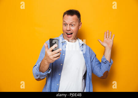 Ritratto di un felice uomo di mezza età guardando il telefono cellulare isolato su sfondo giallo, agitando la mano Foto Stock
