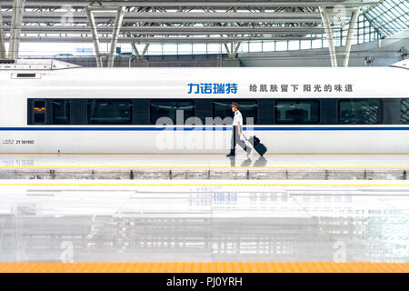 Un asiatico il treno ad alta velocità conduttore passeggiate lungo la piattaforma con un treno auto dietro di lui, mentre si tira la sua valigia in Cina a Shanghai. Foto Stock