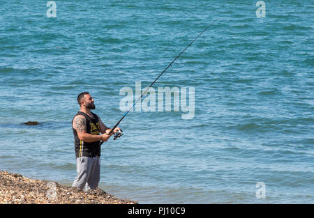 Un uomo in piedi sulla spiaggia vicino al mare in possesso di una canna da pesca la pesca con lenza in mare o il pescatore. Foto Stock