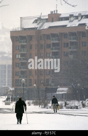 5 Marzo 1993 durante l'assedio di Sarajevo: sul retro del Holiday Inn a livello locale un uomo con una stampella passeggiate attraverso la neve. Un altro giovane uomo porta diverse i contenitori in plastica vuoti, sul suo modo di raccogliere l'acqua. Questo è il territorio da cecchino, ma bella nevicata dà un grado di copertura. Foto Stock