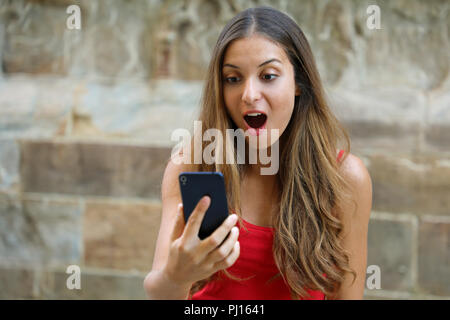 Sorpreso giovane donna utilizzando smart phone all'esterno. Eccitato ragazza urbano utilizzando il nuovo telefono mobile app. Foto Stock