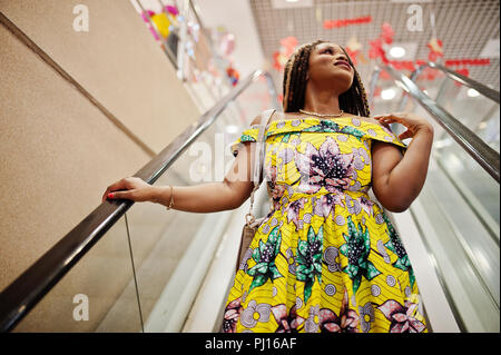 Carino piccola altezza African American Girl con Dreadlocks, usura in corrispondenza di colore giallo, abito in piedi alla scala mobile sul centro commerciale. Foto Stock