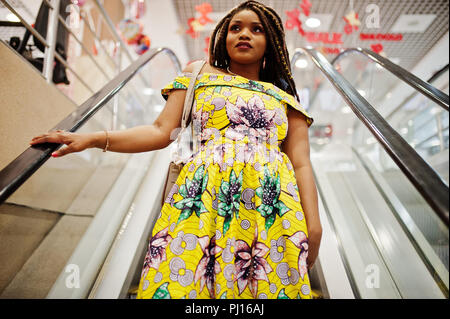 Carino piccola altezza African American Girl con Dreadlocks, usura in corrispondenza di colore giallo, abito in piedi alla scala mobile sul centro commerciale. Foto Stock