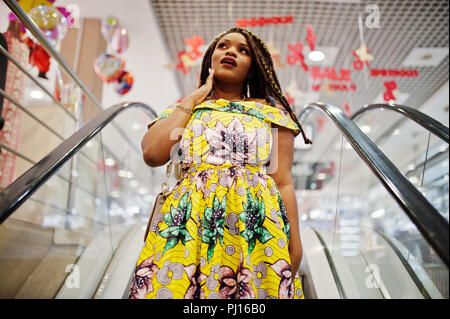 Carino piccola altezza African American Girl con Dreadlocks, usura in corrispondenza di colore giallo, abito in piedi alla scala mobile sul centro commerciale. Sorpresi di fronte. Foto Stock