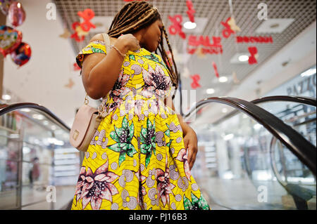 Carino piccola altezza African American Girl con Dreadlocks, usura in corrispondenza di colore giallo, abito in piedi alla scala mobile sul centro commerciale. Foto Stock