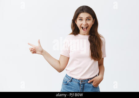 Emozionato e sorpreso moderna moda ragazza con lunghi capelli broadn mascella caduta da stupore che punta a sinistra essendo sotto impressione positiva vedendo incredibile cosa incredibile oltre il muro grigio Foto Stock