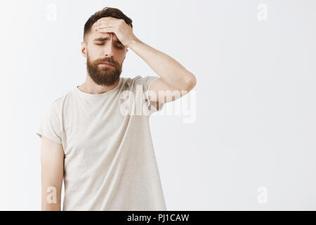 Studio shot dell'uomo sensazione rammarico dopo aver perso durante la competizione sensazione sconvolto e interessati tenendo la mano sulla fronte di chiudere gli occhi e il gemito standing esaurito e stanco oltre il muro grigio Foto Stock
