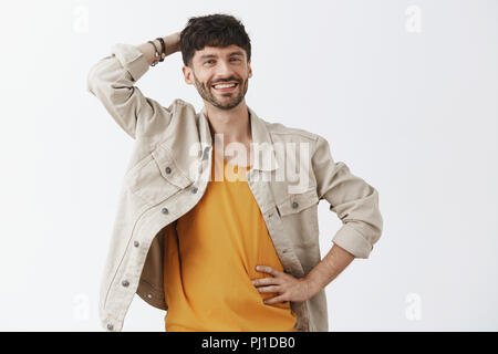 Vita-up shot di carefree cordiale e gioiosa bello amico maschio in camicia al di sopra yellong t-shirt graffiare la testa e tenendo la mano sulla vita sorridente in linea di massima essere rilassati e felici oltre il muro grigio Foto Stock