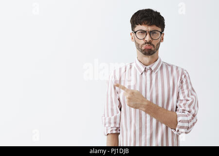 Close-up shot di moody e picky funny guy con la barba e capelli scuri in bicchieri e camicia a righe esercitano le labbra tipo sirena e accigliata da antipatia rivolto in alto a sinistra lamentando Foto Stock