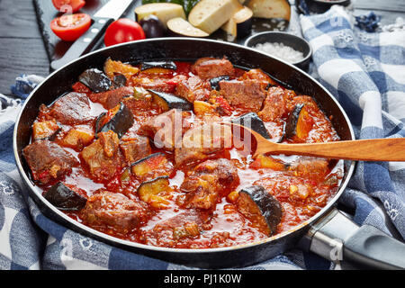 Close-up di melanzane spezzatino di manzo in una padella su tavoli in legno nero con ingredienti e asciugatutto, orizzontale vista da sopra Foto Stock