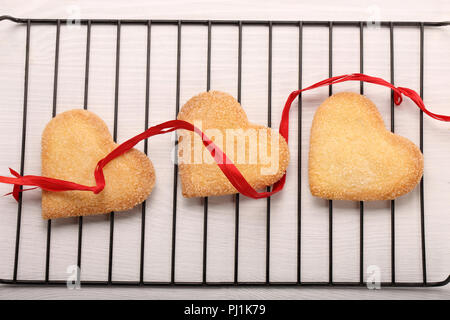 Biscotti croccanti nel cuore sagomato con nastro rosso su bianco sullo sfondo di legno Foto Stock