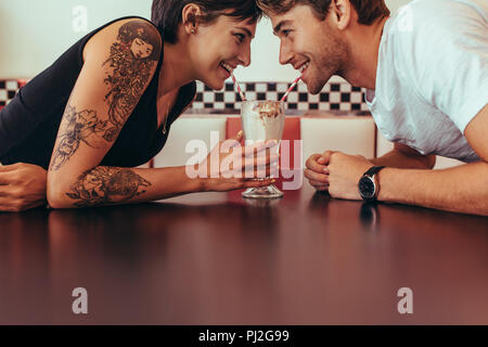 Close up di coppia felice condivisione di un milk shake con cannucce seduto di fronte all'altra in un ristorante. Coppia romantica sorseggiando milk shake da un gl Foto Stock