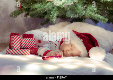 Carino neonato dorme sotto albero di Natale vicino al red doni indossando Santa Claus hat. Foto Stock