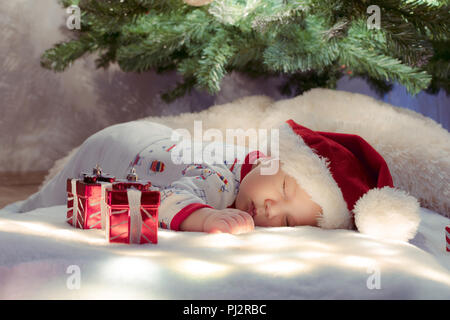 Adorabili neonato dorme sotto albero di Natale nei pressi di doni sulla coperta di illuminazione. Foto Stock