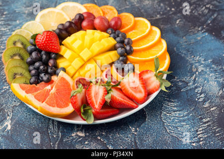 Sani frutti e bacche platter nei colori dell'arcobaleno close up, fragole, mango, uva, arance e kiwi, limoni sul tavolo blu, copiare lo spazio per testo, il fuoco selettivo sul mango Foto Stock