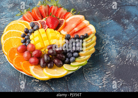 Sani frutti e bacche platter nei colori dell'arcobaleno close up, fragole, mango, uva, arance e kiwi, limoni sul tavolo blu, copiare lo spazio per testo, il fuoco selettivo sul mango Foto Stock