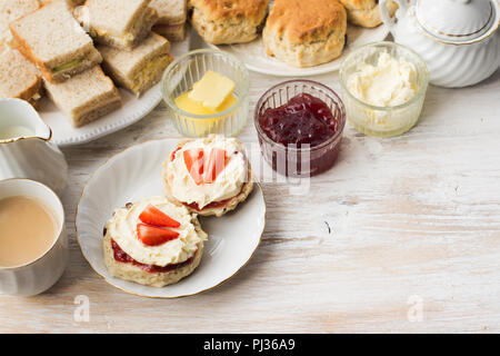 La crema per tè, scones con marmellata e crema di latte, tè con latte, con panini sul retro, sul bianco tavolo in legno, fuoco selettivo copia spazio per il testo Foto Stock