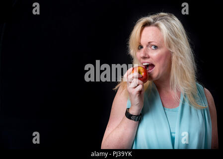 Espressivo donna bionda mordere un apple contro uno sfondo nero Foto Stock