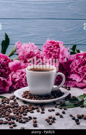 Bella mattina lo sfondo con un caffè in una tazza di chicchi di fiori con le peonie con boccioli di rosa su un calcestruzzo tabella grigia e con un legno blu bac Foto Stock