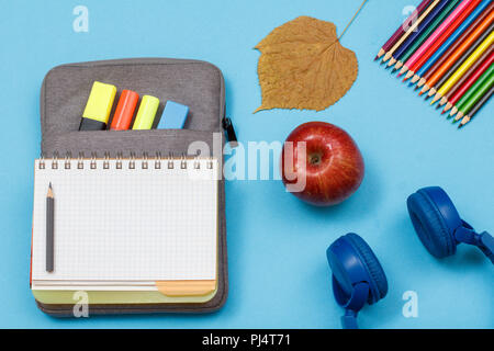 Forniture scolastiche. Aprire il quaderno sul sacchetto custodia a matita con colore pennarelli e marker, foglia secca, matite colorate, apple e cuffie sul backgrou blu Foto Stock