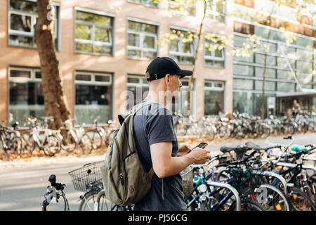 Uno studente con uno zaino o un turista su strada di Lipsia in Germania utilizza un telefono cellulare vicino al parcheggio per le biciclette che si trova accanto alla biblioteca dell'Università di Leipig e ostello della gioventù. Foto Stock