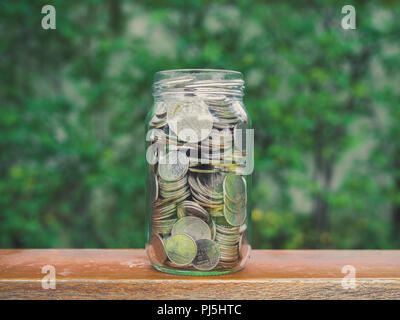 Le monete in un barattolo di vetro sul tavolo in legno e su albero verde dello sfondo. Risparmio di denaro concetto. Foto Stock