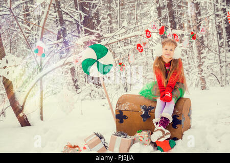 Un po' i capelli rossi ragazza con i capelli lunghi in un lussureggiante mantello e un manto verde con pizzo, in costume del nano assistant Santa Claus in inverno foresta con enormi candy, un petto di doni raccoglie in un cestello e palle di Natale Foto Stock