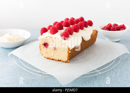 Primo piano di una casalinga lampone focaccia torta su carta pergamena e raffreddamento per rack. Bocce riempito con lamponi freschi e la glassa in background. Foto Stock