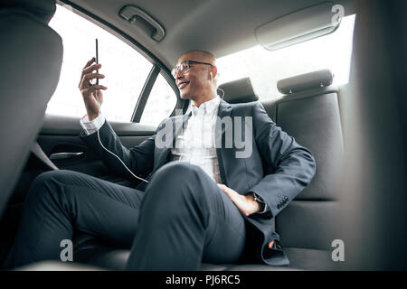 Imprenditore facendo una chiamata video tenendo il suo telefono cellulare mentre è seduto nella sua berlina sul sedile posteriore. Uomo che indossa gli auricolari nella gestione di aziende lavorano su mo Foto Stock