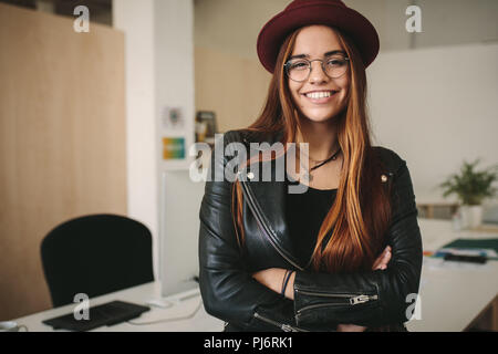 Ritratto di una donna che indossa un cappello e occhiali in piedi in ufficio con bracci incrociati. Sorridente donna di affari in ufficio in piedi con le braccia cros Foto Stock