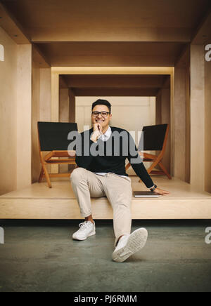 Happy businessman in ufficio seduti in un cubo enclosure con mano sul mento. Imprenditore seduto in ufficio con un computer portatile dal suo lato. Foto Stock