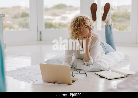 Donna sdraiata sul pavimento e di lavoro sul computer portatile comodamente sdraiato sul pavimento. Donna pensare mentre si lavora sul portatile a casa. Foto Stock
