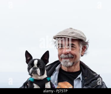 Close up sorridenti senior uomo con le fossette e un graybeard, indossando un cappello, tenendo in mano un simpatico Boston Terrier indossando un cablaggio guardando le camer Foto Stock