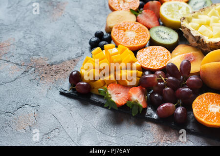 Materie frutti e bacche di piatto sulla pietra grigia sfondo, copia dello spazio. Ananas, mango, satsuma, kiwi, pesche, arance, fragole, uva, blu Foto Stock