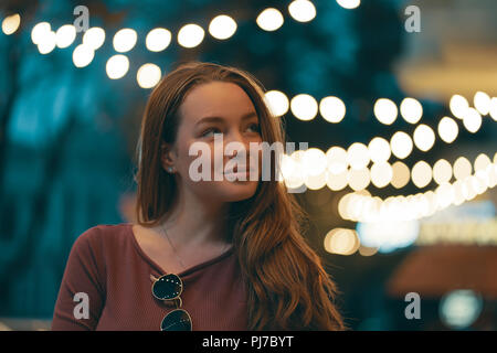 Mezza lunghezza Ritratto di un attraente ragazza vestita in abbigliamento casual con le luci della notte in background. Femmina romantica in piedi in strada illuminat Foto Stock