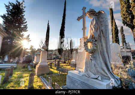 Un angelo di marmo con la sua mano sulla croce, rivolta verso il sole, tra le tombe del cimitero, in concentrazione, tenendo una corona Foto Stock