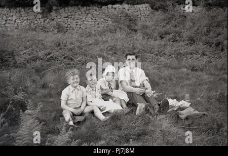 1050s, storico, padre in una camicia e cravatta avente uno spuntino all'aperto in un campo con i suoi tre figli giovani, Inghilterra, Regno Unito. Foto Stock