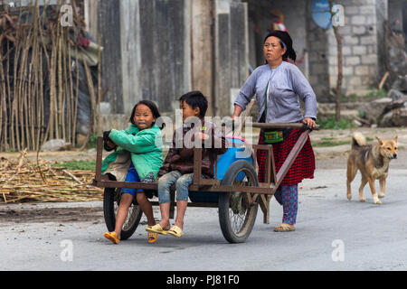 Ha Giang, Vietnam - Marzo 18, 2018: Madre portante due bambini su un carrello manuale in una remota zona rurale del nord del Vietnam Foto Stock