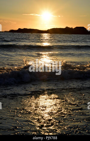 Sunset over Trearddur Bay vicino a Holyhead, Anglesea, il Galles del Nord con la luce del sole che si riflette sull'acqua e onde. Orange sky silhouette seasc paesaggio Foto Stock