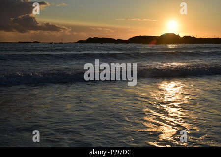 Sunset over Trearddur Bay vicino a Holyhead, Anglesea, il Galles del Nord con la luce del sole che si riflette sull'acqua e onde. Orange sky silhouette seasc paesaggio Foto Stock