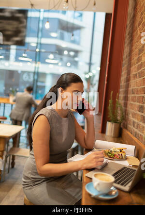 Imprenditrice parlando su smart phone, lavorando al computer portatile in cafe Foto Stock