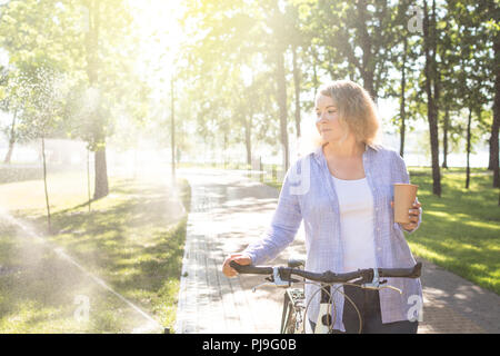 Giovane donna blonge camminando la sua bicicletta nel parco e tenendo una tazza di caffè in mano. Foto Stock
