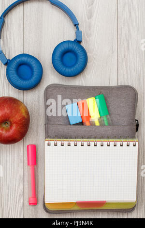 Forniture scolastiche. Aprire il quaderno sul sacchetto custodia a matita con colore pennarelli e marker, Apple, cuffie su grigio di tavole di legno. Vista da sopra con copia sp Foto Stock