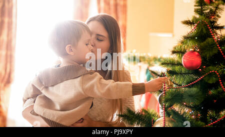 Bella giovane donna baciare il suo bambino mentre egli è la decorazione di albero di Natale Foto Stock