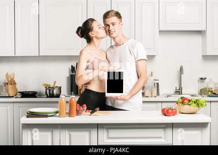 Ragazza baciare ragazzo ed egli holding tablet con schermo vuoto in cucina Foto Stock