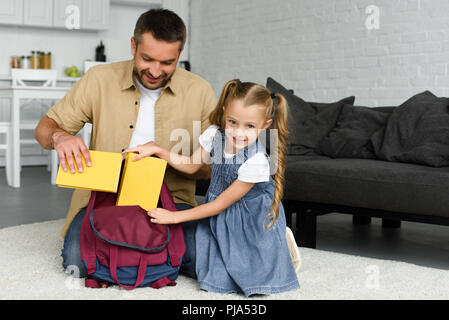 Padre sorridente aiutando piccola figlia imballaggio zaino per il primo giorno di scuola, torna al concetto di scuola Foto Stock